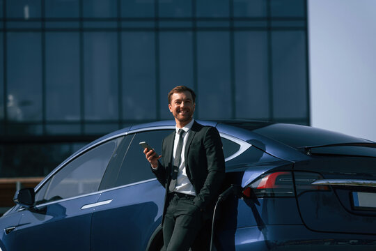 Smiling, holding smartphone. Businessman is standing near his electric car outdoors