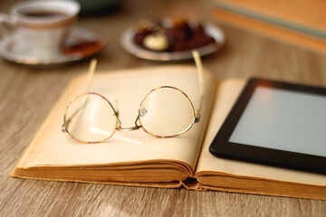 Books, reading glasses, e-reader, plate of chocolate pralines, bowl of cookies, cups of tea and lit candles on the table. Selective focus.