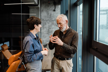 Smiling mature colleagues discussing work while standing near office window
