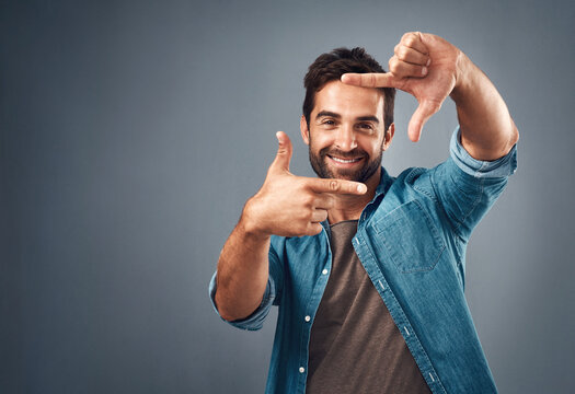 Frame, hands and portrait of man in studio on gray background with happiness, confident and smile. Finger border, mockup and male person face with hand sign for picture, photography and perspective