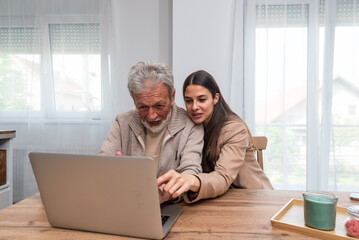 Daughter and elderly father or grandfather sitting at the table at home daughter teaches him to use laptop computer and surf the internet to find information easily entertainment and read news