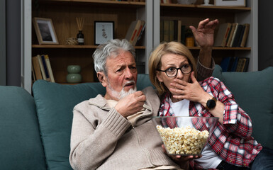 Elderly couple watching bad news, quiz or competition with displeasure and expression while eating popcorn. Senior people husband and wife watch political debate at home making negative faces