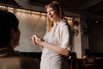 Positive waitress writes down order of mature woman at table in cafe