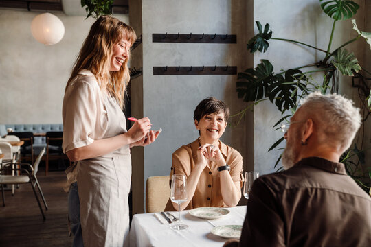 Laughing young blonde waitress writes down order of elderly couple in cafe