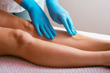 Hands of a cosmetologist close-up, in blue gloves, applying sugar paste on a woman's leg. Depilation procedure