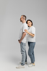 full length of tattooed woman with short brunette hair leaning on back of smiling husband while standing together in white t-shirts and denim jeans on grey background, happy couple