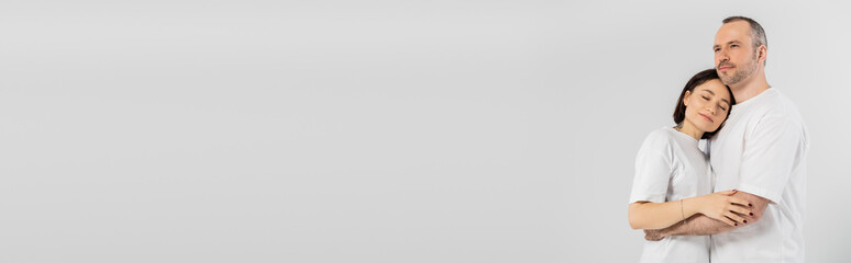 tattooed woman with short brunette hair leaning on chest of husband while standing together in white t-shirts isolated on grey background, happy couple, banner