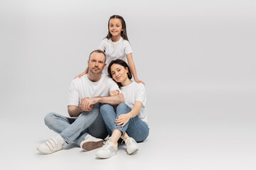 joyous preteen girl hugging mother and father in white t-shirts and blue denim jeans while bonding together and looking at camera on grey background, Happy children's day