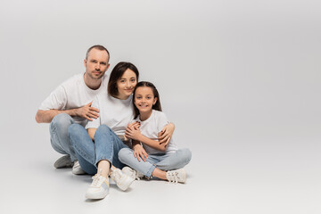 joyous father and mother with short hair hugging cheerful preteen daughter while sitting with crossed legs in white t-shirts and blue denim jeans on grey background, Happy children's day