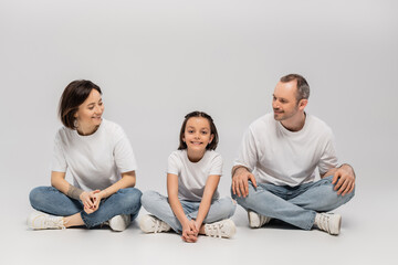 Fototapeta na wymiar joyous father and tattooed mother with short hair looking at cheerful preteen daughter while sitting with crossed legs in white t-shirts and blue denim jeans on grey background, Happy children's day