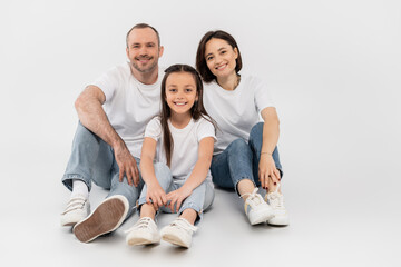 stylish family in white t-shirts and blue denim jeans looking at camera and sitting together on grey background, International child protection day, happy parents and daughter