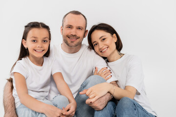 portrait of happy family in white t-shirts and blue denim jeans looking at camera and sitting together on grey background, International child protection day, parents and daughter