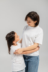 happy mother with short hair and tattoo on hand hugging brunette daughter while standing together in white t-shirts and blue denim jeans on grey background, International child protection day