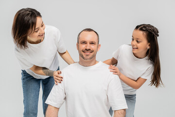 woman with short hair and tattoo on hand and brunette teenage girl in blue denim jeans looking at man in white t-shirt on grey background, happy family