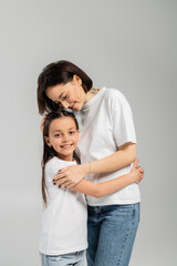 Fototapeta premium happy mother with short hair and tattoo on hand hugging preteen daughter while standing together in white t-shirts and blue denim jeans on grey background, International child protection day