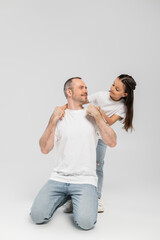happy girl with long brunette hair leaning on unshaved and cheerful father with bristle while posing in white t-shirts and denim jeans while looking at each other on grey background