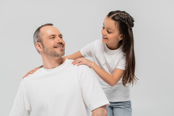 happy preteen girl with long brunette hair leaning on unshaved and cheerful father with bristle while posing in white t-shirts and looking at each other on grey background, Child protection day