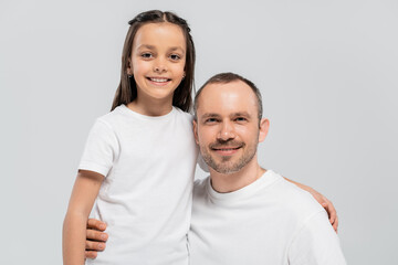 Fototapeta premium unshaved and cheerful father with bristle hugging happy preteen daughter with long brunette hair while posing in white t-shirts and looking at camera on grey background, Child protection day