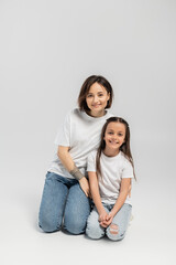 happy mother with tattoo on hand and short hair hugging preteen brunette daughter while sitting together in white t-shirts and blue denim jeans on grey background, child protection day