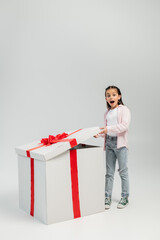 Full length of shocked preteen girl in casual clothes looking at camera while opening big gift box during child protection day celebration on grey background