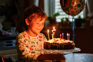 Adorable happy blond little kid boy celebrating his birthday. Child blowing seven candles on homemade baked cake, indoor. Birthday party for school children, family celebration of 7 years