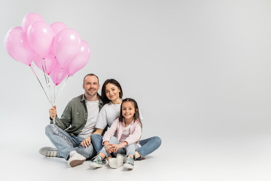 Smiling parents in casual clothes hugging preteen daughter and holding festive pink balloons while celebrating international children day on grey background
