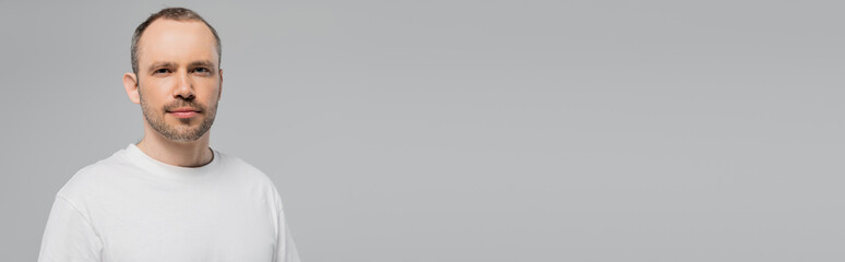 Fototapeta premium unshaved man with bristle standing in white t-shirt and looking at camera while posing isolated on grey background in studio, copy space, confidence and masculinity, banner