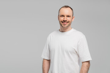 Fototapeta premium unshaved happy man with bristle standing in white t-shirt and looking at camera while posing isolated on grey background in studio, copy space, happiness