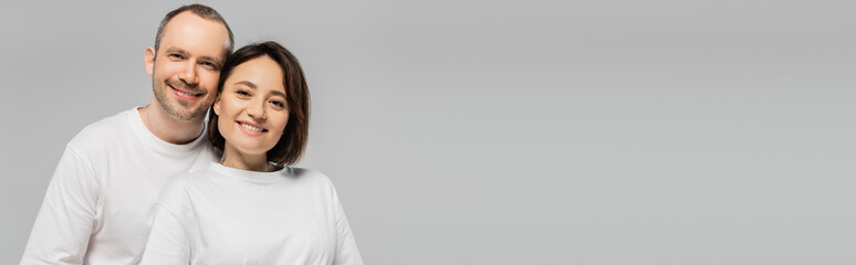 positive husband and tattooed wife with short brunette hair standing together in white t-shirts and looking at camera isolated on grey background in studio, happy couple, banner