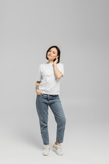 Fototapeta na wymiar full length of appealing and tattooed woman with short hair and natural makeup standing in white t-shirt and posing with hand in pocket of blue denim jeans while looking at camera on grey background