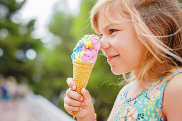 Happy preschool girl eating colorful ice cream in waffle cone on sunny summer day. Little toddler...