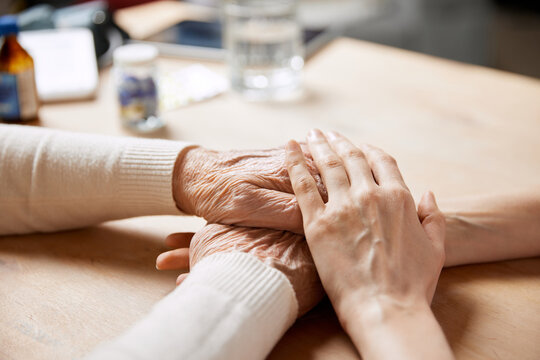 Close-up image of doctor's hand holding senior female patient hands, giving support and hope. Table with pills and meds. Concept of medical care, medicine, illness, health care, profession