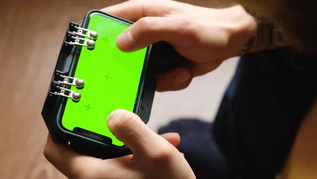 Caucasian man using smartphone in horizontal mode with green mock-up screen sits on yellow bed and plays online games. Guy uses joystick for phone. Focus on Chroma Display. View from above.
