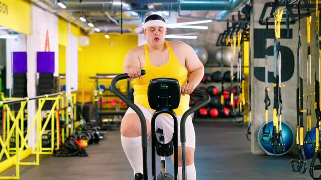 The fat man is exercising in the gym and looking at the burger and pizza. Obesity of a young man and doing sports in the gym. The fat man eats a burger.