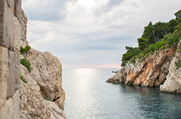 Fototapeta na wymiar Landscape scenery of coastal rock under forest, calm sea water and stormy clouds in the horizon. Summer beauty of adriatic seaside nature.