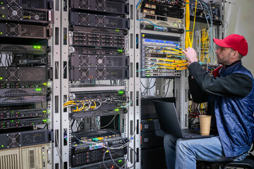 A man with a laptop sits on a chair in a data center. The system administrator works in the server room.A technician connects wires to a central router.