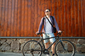Happy young caucasian man in casual street style standing with bike against wall on street and...