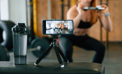 Doing squats, shooting video by smartphone. Beautiful strong woman is in the gym