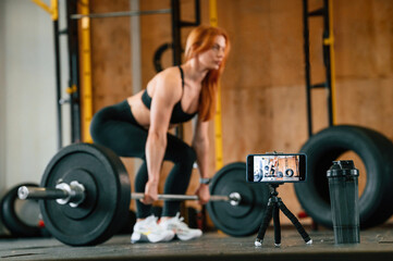 With barbell. Shooting video by smartphone. Beautiful strong woman is in the gym