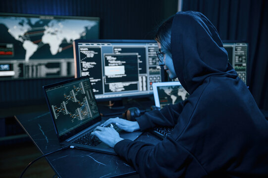 Black colored hoodie. Young professional female hacker is indoors by computer with lot of information on displays