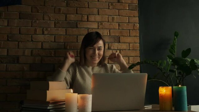 Happy caucasian woman using laptop at home during electricity outage or blackout with lit candles. Inspired woman raises her hands up after goal achievement. Success concept