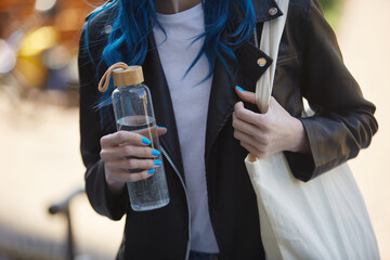 Young woman holding a reusable glass bottle of water and wearing a eco shopper bag on shoulder. Responsible female person leading a sustainable lifestyle