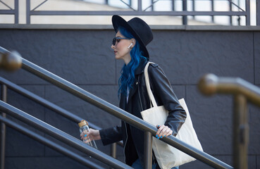Diverse female model with dyed blue hair walking on stairs in the city center wearing a cotton tote bag on shoulder and holding a glass water bottle in hand