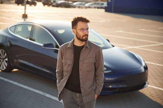 In glasses. Man is standing near his electric car outdoors