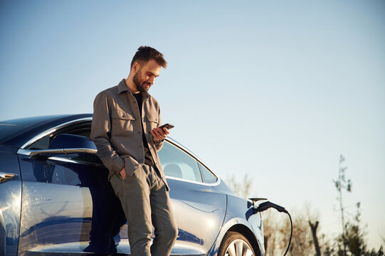 Holding smartphone in hand. Man is standing near his electric car outdoors - Powered by Adobe