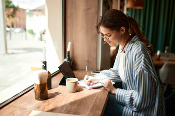 Fototapeta premium Portrait with concentrated young girl, student writing in notebook while learning with tablet and coffee cup in cafe. Freelance work, business people