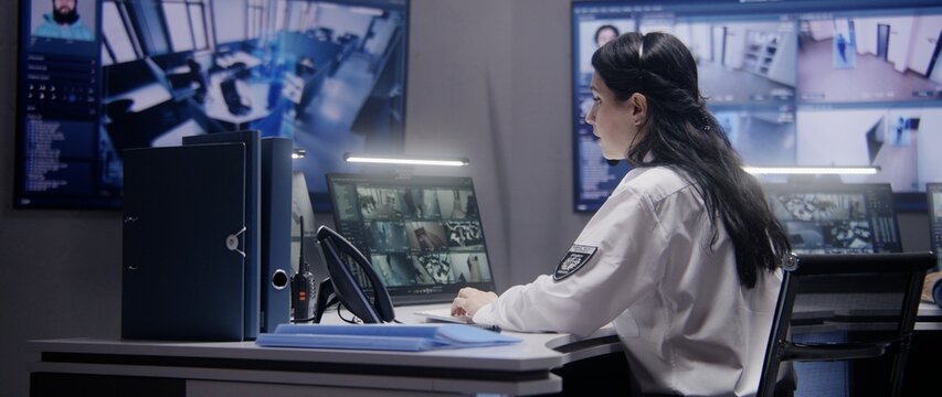 Female security officer works on computer in police monitoring center. Multi ethnic workers control surveillance CCTV cameras with tracking AI face recognition system on tablet and big digital screen.