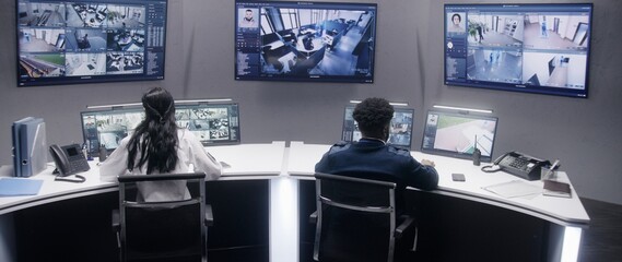 Multi ethnic security officers monitor CCTV cameras with AI face scanning. Multiple big screens...