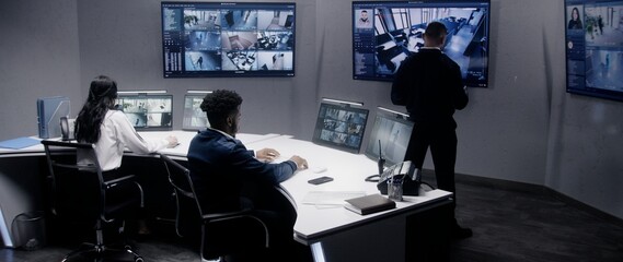 Multi ethnic security officers monitor CCTV cameras with AI face scanning. Multiple big screens with displayed security cameras. Teamwork in surveillance room. Monitoring, tracking and social safety. - obrazy, fototapety, plakaty