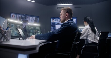 Male security officer touches tablet and controls CCTV cameras with AI face scanning. Computer monitors and big digital screens showing security cameras footage. Female colleague works at background.